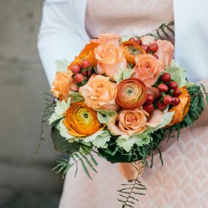 Woman holding a bouquet of orange garden roses and ranunculus flowers decorated with greenery.